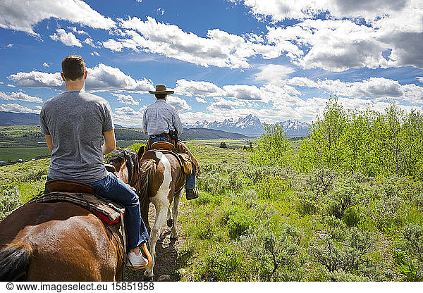 Two men ride horses with the Teton mountain range in the distance