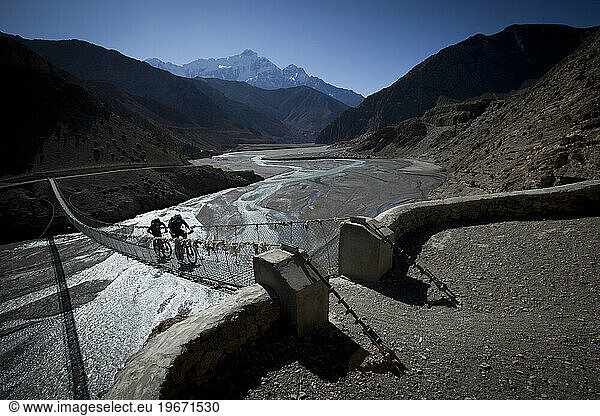 Two men ride mountain bikes across small bridge over river valley with mountains in background.