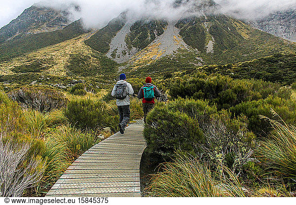 Two men walking on wooden path toward mountains in the clouds ahead.