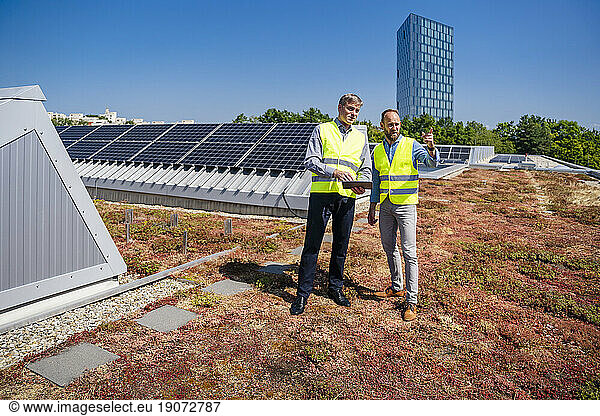 Two men with tablet PC wearing reflective vests talking on the roof of a company building with solar panels