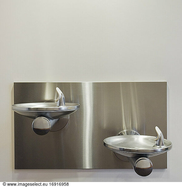 Two metal drinking fountains mounted on wall at different heights.