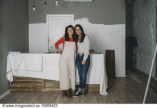 Two mid adult women leaning against table while painting in their new shop  full length portrait