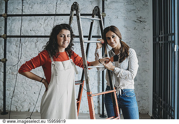 Two mid adult women leaning on step ladders with paint brush in their new shop  portrait