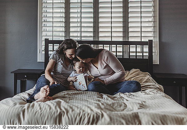 Two moms reading with baby while snuggling on bed