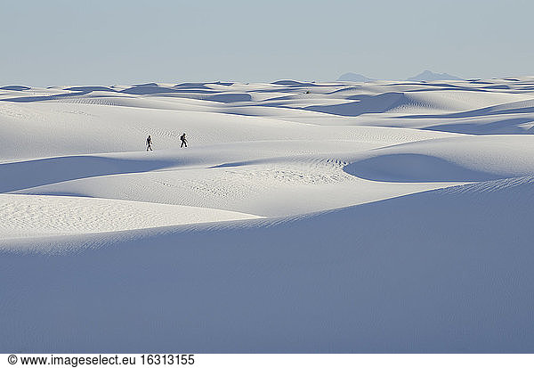 Two people at a distance walking across white sand dunes.