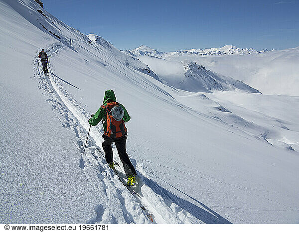 Two people backcountry skiing up a track through the snow on a sunny day.