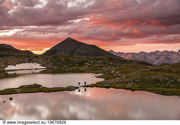 Two people hike along the edge of a lake in the mountains at sunset.