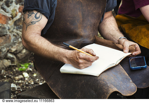 Two people  man and woman blacksmiths wearing leather aprons writing into a notebook sat in a garden.