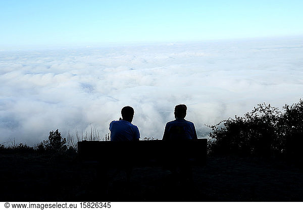two people sitting on bench on mountain top overlooking clouds