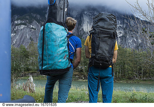Two people with big backpack standing in front of mountain and river