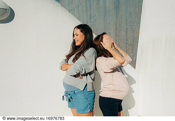 Two pregnant women standing against wall during sunny summer day
