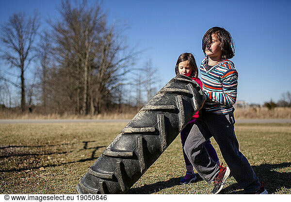 two small children work together to lift a heavy tire in a park