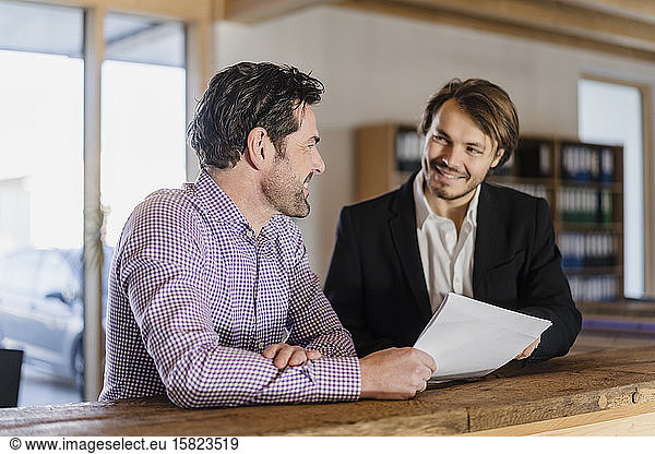 Two smiling businessmen with documents talking in wooden open-plan office