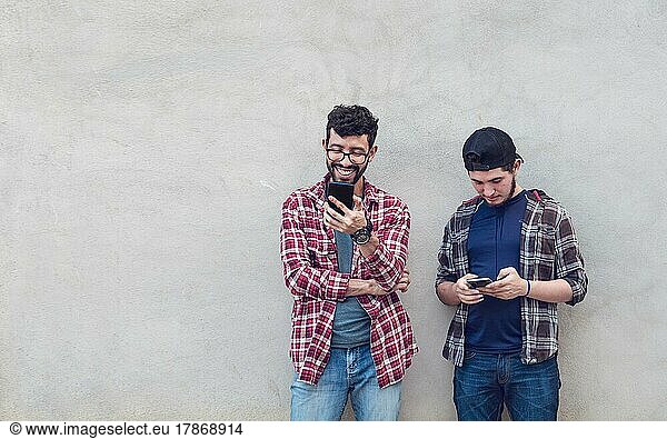 Two smiling friends leaning on a wall checking their cell phones  Friends leaning on a wall texting on their phones. Friend showing cell phone to his friend  Smiling friends checking cell phones