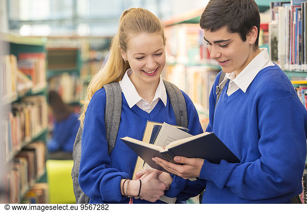 Two smiling high school students wearing school uniforms looking at book in library