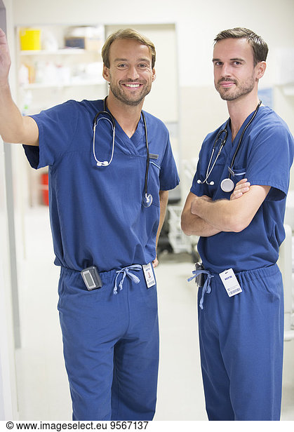 Two smiling male doctors wearing scrubs standing in hospital ward