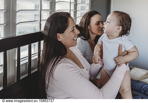 Two smiling moms holding baby daughter at home near window