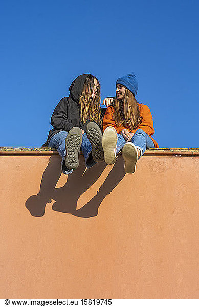 Two smiling teenage girls sitting on a wall against blue sky