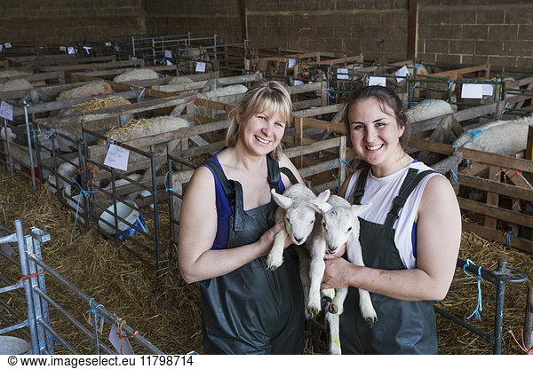 Two smiling women standing in a stable  holding newborn lambs.
