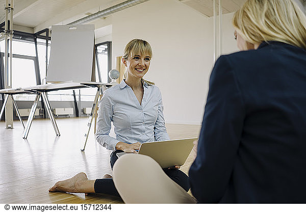 Two smiling young businesswomen sitting on the floor and talking in office