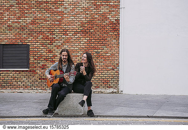 Two smiling young musicians sitting on a stone playing guitar