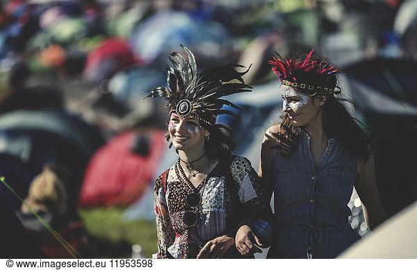Two smiling young women at a summer music festival face painted  wearing feather headdress  standing among tents.