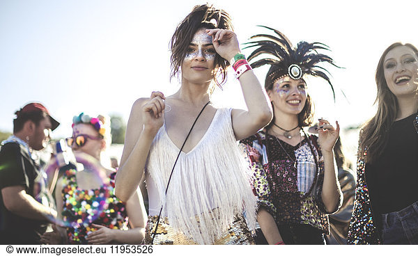 Two smiling young women at a summer music festival face painted  wearing feather headdress  standing among the crowd  looking at camera.