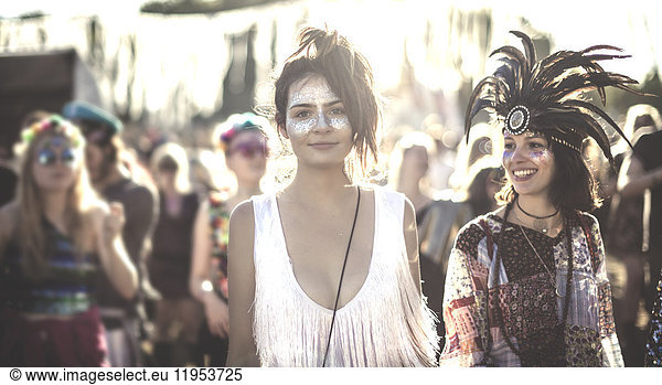 Two smiling young women at a summer music festival face painted  wearing feather headdress  standing among the crowd  looking at camera.