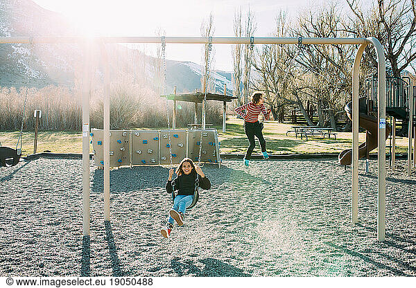 Two teen girls swing on swings at playground with snow in background
