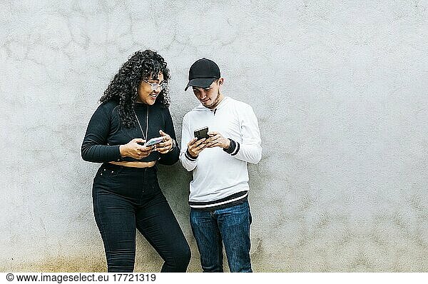Two teenage friends checking their cell phones and smiling  Two teenagers together checking their cell phones  Guy and girl leaning on a wall checking their cell phones  Two smiling friends checking their cell phones