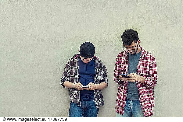 Two teenage friends on a wall checking their cell phones  Two friends leaning on a wall texting on their phones. Friend showing cell phone to his friend  Smiling friends checking cell phones