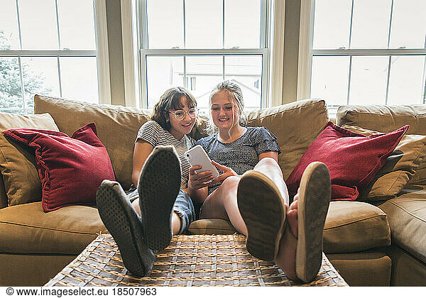 Two teenage girls sitting on couch with feet up looking at cellphone.