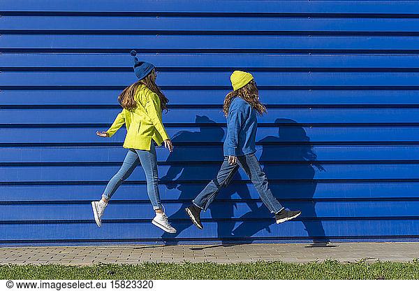 Two teenage girls wearing matching clothes jumping in the air in front of blue background