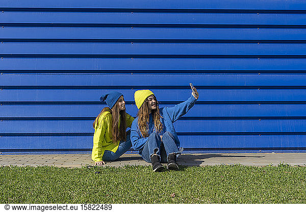 Two teenage girls wearing matching clothes taking selfie with smartphone in front of blue background
