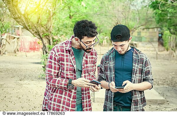 Two teenage guys pointing and checking their cell phones  Two friends looking at the content of their cell phone. Friend showing cell phone to his friend  Smiling friends checking cell phones