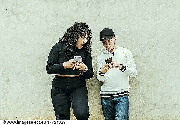 Two teenagers together checking their cell phones  Two smiling friends checking their cell phones  Two teenage friends checking their cell phones and smiling  Guy and girl leaning on a wall checking their cell phones