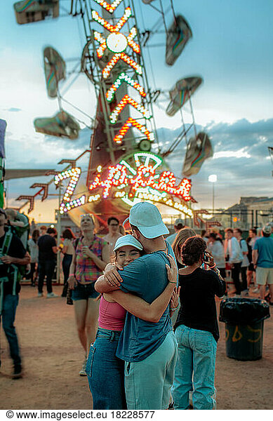 Two teens hugging in front of a carnival ride at sunset