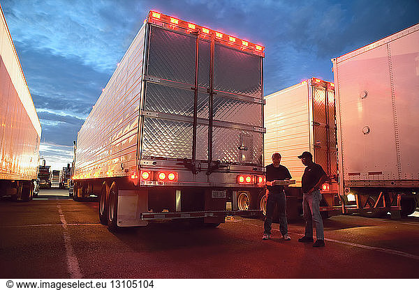 Two truck drives checking dispatch papers while standing next to truck trailers in a large parking lot at night.