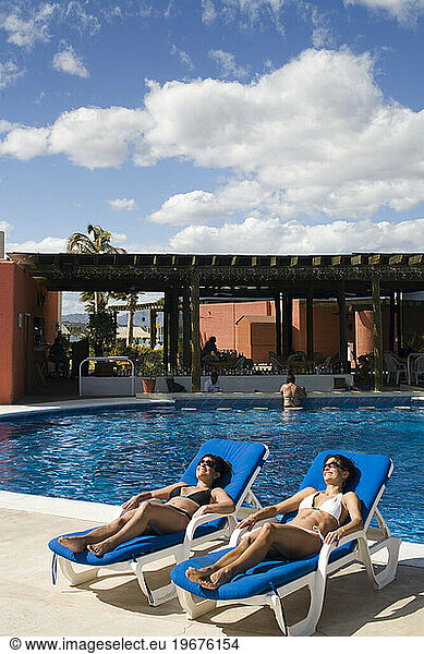 Two women sit near a pool at a hotel underneath a partly cloudy sky  Loreto  Baja California Sur  Mexico.