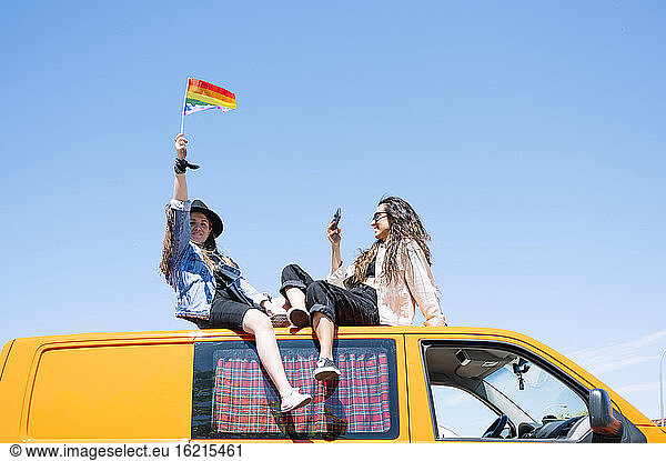 Two women sitting on roof of van  waving pride flag