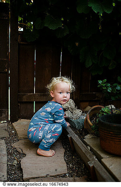 Two Year Old Boy in Pajamas Crouching in Front Yard Under Grapevine