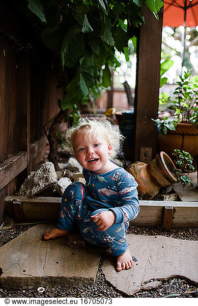 Two Year Old Boy in Pajamas Sitting on Rocks Under Grapevine in Yard