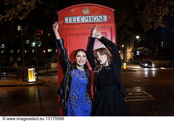 Two young female friends dancing in front of red phone box at night  London  UK