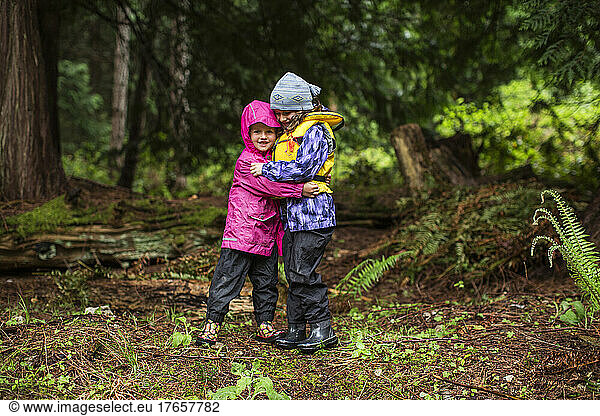 Two young girls in rain gear stand and hug in a forest