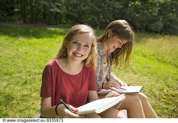 Two young girls sitting outside on the grass  with sketch pads and pencils.