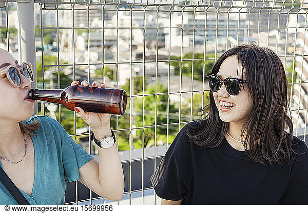 Two young Japanese women sitting on a rooftop in an urban setting  drinking beer.