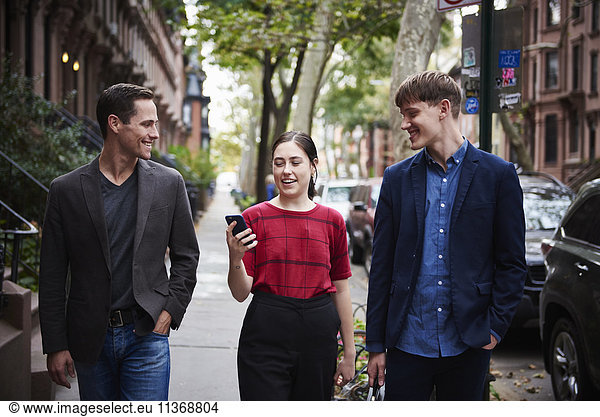 Two young men and a young woman walking down a city street looking at a cellphone.