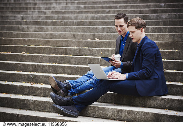 Two young men sitting on a flight of steps looking at a laptop together.