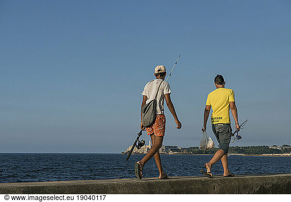 Two young men walking along the top of the MalecÃ?Â³n (Avenida de Maceo)  carrying fishing rods. Havana  La Habana  Cuba.