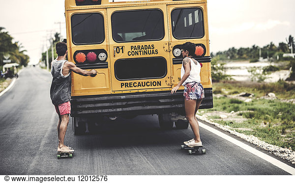 Two young people skateboarding while holding on to a moving school bus.